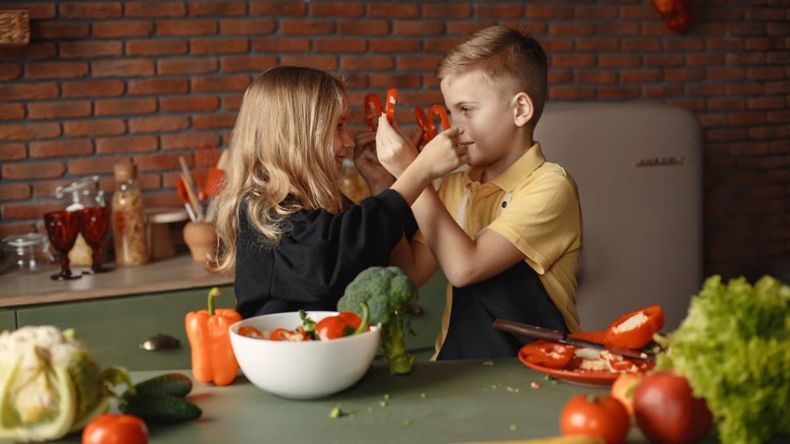 Playful boy and girl wearing apron preparing healthy vegetarian salad together at table with ingredients and bowls while looking at each other through slices of pepper