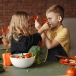Playful boy and girl wearing apron preparing healthy vegetarian salad together at table with ingredients and bowls while looking at each other through slices of pepper