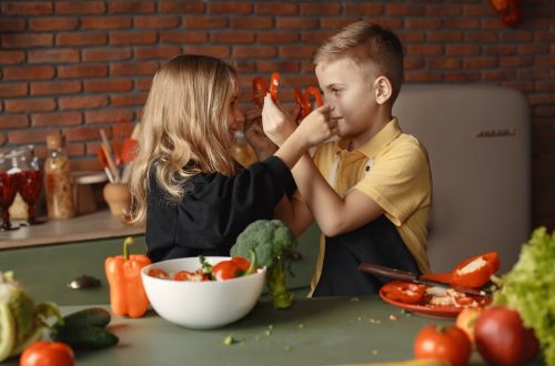 Playful boy and girl wearing apron preparing healthy vegetarian salad together at table with ingredients and bowls while looking at each other through slices of pepper