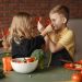 Playful boy and girl wearing apron preparing healthy vegetarian salad together at table with ingredients and bowls while looking at each other through slices of pepper